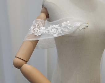 Puede incluir: Una manga blanca y transparente, con hombros descubiertos y detalles de encaje floral. La manga está sujeta a un maniquí, mostrando el delicado diseño y la textura de la tela. El encaje presenta intrincados patrones florales.