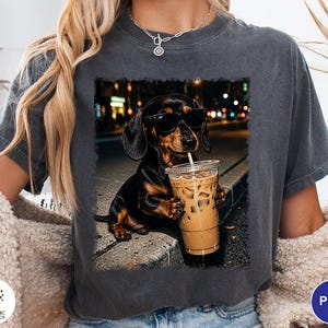 May include: A charcoal gray t-shirt featuring a graphic print of a black and tan dachshund wearing black sunglasses and holding a clear plastic cup filled with iced coffee, set against a blurred nighttime city street background.