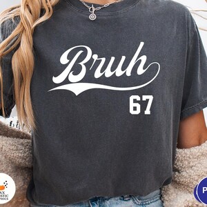 May include: Dark gray t-shirt with the word "Bruh" in a white script font and the number "67" below it. The shirt has a vintage, slightly faded look.