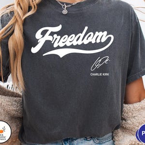 May include: Dark gray t-shirt with the word "Freedom" in white script. Below the word is a signature and the name "Charlie Kirk". The shirt is a casual style.