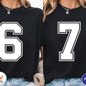 Op de afbeelding: Twee zwarte sweatshirts, elk met een groot wit nummer. E&eacute;n toont het nummer 6, de andere het nummer 7. Beide nummers hebben een vetgedrukt, omlijnd ontwerp. De sweatshirts worden door een persoon gedragen, met een zichtbare ketting.