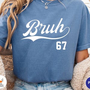 May include: A blue t-shirt with the word "Bruh" in a white script font, with the number "67" below it. The shirt is a solid color and has short sleeves.