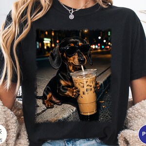 May include: A black t-shirt featuring a graphic print of a black and tan dachshund wearing black sunglasses and holding a clear plastic cup filled with iced coffee. The dog is positioned on a concrete curb with a blurred city street background.
