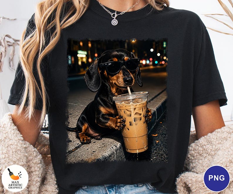 May include: A black t-shirt featuring a graphic print of a black and tan dachshund wearing black sunglasses and holding a clear plastic cup filled with iced coffee. The dog is positioned on a concrete curb with a blurred city street background.