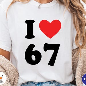 Op de afbeelding: Wit T-shirt met de tekst "I &hearts; 67" in zwart en rood. Het hartsymbool is rood. De cijfers "67" zijn vetgedrukt, zwart. Het shirt is een casual stijl.
