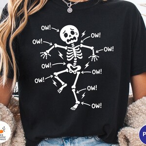May include: Black t-shirt featuring a white skeleton graphic with the word "OW!" and lightning bolts pointing to various body parts. The skeleton has a distressed expression, conveying pain. A fun, spooky design.