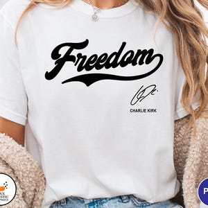 May include: White t-shirt with the word "Freedom" in a black script font. Below the word is a signature and the name "Charlie Kirk" in a smaller font. The shirt is a casual style.