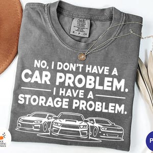 May include: A gray Comfort Colors t-shirt with the text "NO, I DON'T HAVE A CAR PROBLEM. I HAVE A STORAGE PROBLEM." and three car illustrations. A gold necklace and a brown hat are also visible.