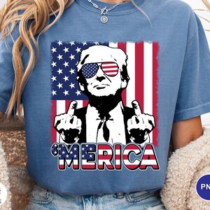 May include: Blue t-shirt featuring a graphic of a man wearing sunglasses with an American flag design. The image includes the text "'MERICA" in red, white, and blue. The man is giving the middle finger with both hands.