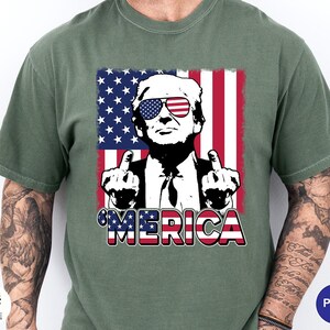 May include: Green t-shirt featuring a graphic of a man wearing sunglasses with an American flag design. The image includes the word "'MERICA" in red, white, and blue. The man is giving the middle finger with both hands.