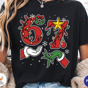 May include: Black t-shirt with a festive Christmas design. The design features the numbers 67 in red, adorned with Christmas lights, a star, and ornaments. Includes a Santa hand and a Grinch hand. Snowflakes and stars complete the holiday theme.