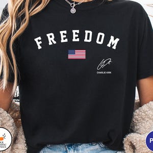 May include: Black t-shirt with the word "FREEDOM" in white arched letters above a small American flag. Below the flag is a signature and the name "CHARLIE KIRK".