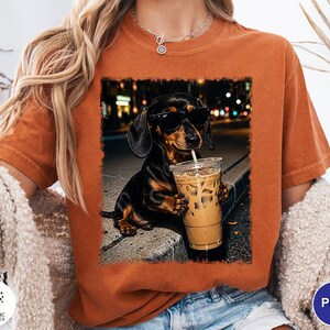 May include: A burnt orange t-shirt featuring a graphic print of a black and tan dachshund wearing black sunglasses and holding a clear plastic cup filled with iced coffee. The dog is positioned on a curb with a blurred city street background.