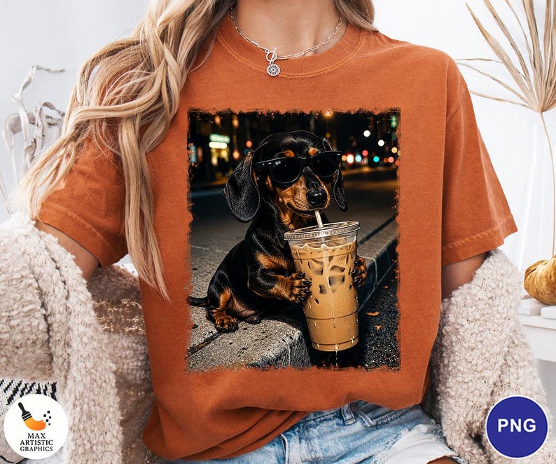 May include: A burnt orange t-shirt featuring a graphic print of a black and tan dachshund wearing black sunglasses and holding a clear plastic cup filled with iced coffee. The dog is positioned on a curb with a blurred city street background.