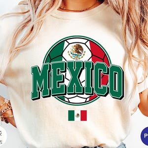 May include: An ivory-colored t-shirt featuring a large graphic of a soccer ball colored with the green, white, and red stripes of the Mexican flag. The word MEXICO is printed in large, bold green letters across the center. A small Mexican flag is below.