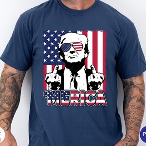May include: Navy blue t-shirt featuring a graphic of a person wearing sunglasses with an American flag design. The person is giving the middle finger. The word "'MERICA" is printed in red, white, and blue.