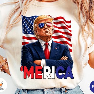 May include: Cream-colored sweatshirt featuring a graphic of a man wearing sunglasses with an American flag design. The man is in a suit with a red tie, and the word "'MERICA" is printed below. An American flag is in the background.