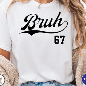 May include: White t-shirt with the word "Bruh" in a black script font above the number "67". The shirt is a casual style, suitable for everyday wear.