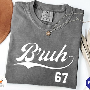 May include: A gray Comfort Colors t-shirt with the word "Bruh" in white script and the number "67" below. A gold necklace with a pendant is draped on the shirt. The shirt is folded, with a brown hat and dried leaves in the background.