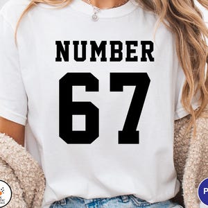 Op de afbeelding: Wit T-shirt met het woord "NUMBER" in dikke zwarte letters boven het nummer "67" in een groot, blokkerig lettertype. Het shirt is effen en eenvoudig, geschikt voor vrijetijdskleding.