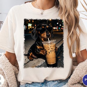 May include: A cream-colored t-shirt featuring a graphic print of a black and tan dachshund wearing black sunglasses and holding a clear plastic cup filled with iced coffee. The dog is positioned on a curb with a blurred city street background.