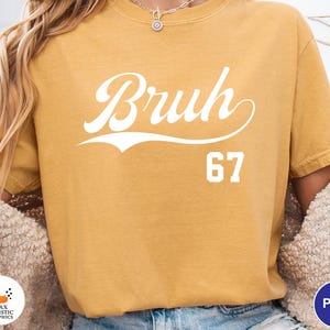May include: Mustard yellow t-shirt with the word "Bruh" in white script, above the number "67". The shirt has a vintage, slightly faded look. The design is centered on the front of the shirt.