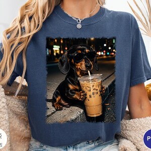 May include: A dark blue short-sleeve t-shirt featuring a graphic print of a black and tan dachshund wearing black sunglasses and holding a clear plastic cup filled with iced coffee, set against a blurred city street background at night.