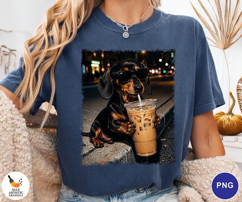 May include: A dark blue short-sleeve t-shirt featuring a graphic print of a black and tan dachshund wearing black sunglasses and holding a clear plastic cup filled with iced coffee, set against a blurred city street background at night.