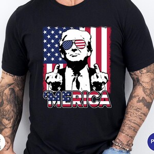 May include: Black t-shirt featuring a graphic of a man wearing sunglasses with the American flag design. The man is giving the middle finger with both hands. The word "MERICA" is written in red, white, and blue.