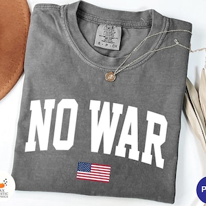 以下が含まれることがあります： 「NO WAR」の文字が大きく白でプリントされたグレーのTシャツ。テキストの下には小さなアメリカ国旗があります。ペンダント付きのゴールドネックレスがシャツの上に置かれています。