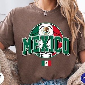 May include: A brown short-sleeve t-shirt featuring a graphic of a soccer ball colored with the green, white, and red of the Mexican flag. The word MEXICO is printed in large green letters with a white outline. A small Mexican flag is below the ball.