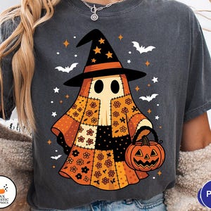 May include: Dark gray t-shirt featuring a Halloween ghost character wearing a witch hat and holding a pumpkin basket. The ghost has a patchwork design in orange, black, and cream colors, with stars and bats scattered around the design.