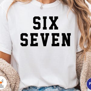 Op de afbeelding: Wit T-shirt met de woorden "SIX SEVEN" in vette, zwarte blokletters. Het shirt heeft een eenvoudig ontwerp met een ronde hals. De tekst is gecentreerd op de voorkant van het kledingstuk.