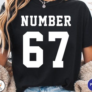 Op de afbeelding: Zwart T-shirt met het woord "NUMBER" in witte blokletters boven het witte nummer "67". Het shirt heeft een ronde hals. Er is een klein logo zichtbaar in de linker benedenhoek.