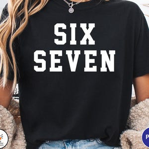 Op de afbeelding: Zwart T-shirt met de woorden "SIX SEVEN" in grote, witte blokletters. Het shirt heeft een ronde hals. Het model draagt een cr&egrave;me-kleurige trui.