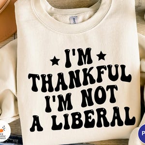 May include: Cream-colored sweatshirt with the text "I'M THANKFUL I'M NOT A LIBERAL" in black, retro-style font. The design includes stars above the words "I'M" and a small logo in the bottom left corner.