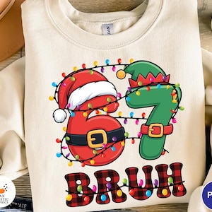 May include: Cream-colored sweatshirt with a festive Christmas design. The graphic features the numbers "67" decorated with a Santa hat and elf hat, strung with colorful lights, and the word "BRUH" in a plaid pattern.