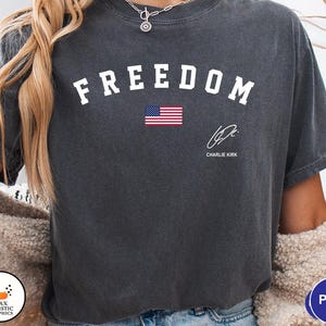 May include: Dark gray t-shirt with the word "FREEDOM" in white block letters above a small American flag. Below the flag is a signature and the name "CHARLIE KIRK".