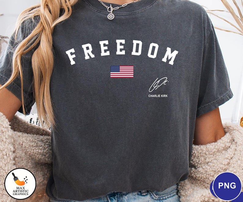 May include: Dark gray t-shirt with the word "FREEDOM" in white block letters above a small American flag. Below the flag is a signature and the name "CHARLIE KIRK".