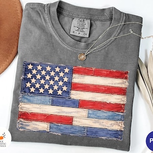 May include: A grey Comfort Colors t-shirt with a distressed American flag design. The flag is made of red, white, and blue patches with white stars. A silver necklace with a small pendant rests on the shirt.
