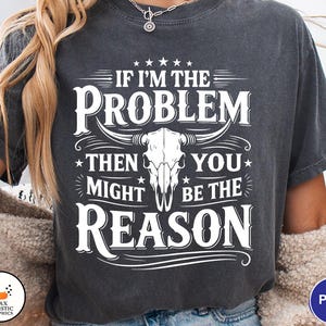 Può includere: T-shirt grigio scuro con la scritta bianca "IF I'M THE PROBLEM THEN YOU MIGHT BE THE REASON", con grafica di un teschio di toro. La maglietta ha un aspetto vintage e usurato.