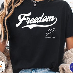 May include: Black t-shirt with the word "Freedom" in a white script font. Below the word is a signature and the name "Charlie Kirk" in a smaller font. The shirt is a solid black color.