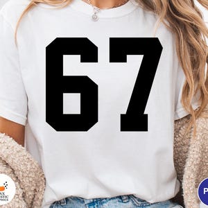 Op de afbeelding: Wit T-shirt met het zwarte nummer 67 in een vetgedrukte blokletter. Het shirt wordt door een persoon gedragen. De afbeelding toont de bovenste helft van de persoon die het shirt draagt.