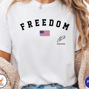 May include: White t-shirt with the word "FREEDOM" in black arched lettering above a small American flag graphic. Below the flag is a signature and the name "Charlie Kirk".