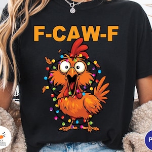 Puede incluir: Camiseta negra con el texto "F-CAW-F" en naranja sobre un dibujo animado de un pollo. El pollo es naranja con una cresta roja y está decorado con luces navideñas de colores, con una expresión de sorpresa.