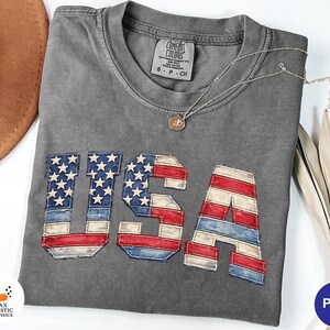 Puede incluir: Camiseta gris Comfort Colors con un diseño desgastado de la bandera de EE. UU. Las letras 'USA' son rojas, blancas y azules, con un aspecto vintage. Un collar plateado con un pequeño colgante descansa sobre la camiseta. Talla S-P-CH.