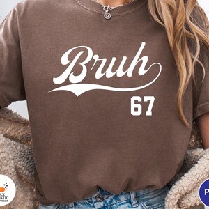 May include: A brown t-shirt with the word "Bruh" in white script and the number "67" below it. The shirt has a vintage, worn look.
