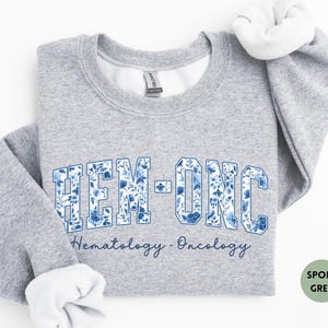 May include: Sport grey sweatshirt with the text "HEM-ONC" in a blue floral design. Below the text is the phrase "Hematology - Oncology" in a script font. The sweatshirt has white lining at the collar and cuffs.