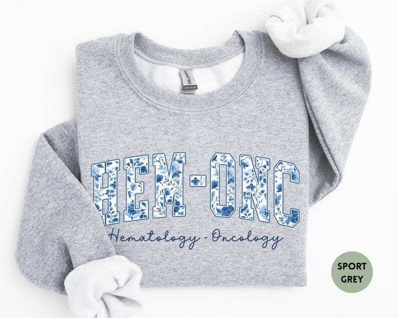 May include: Sport grey sweatshirt with the text "HEM-ONC" in a blue floral design. Below the text is the phrase "Hematology - Oncology" in a script font. The sweatshirt has white lining at the collar and cuffs.