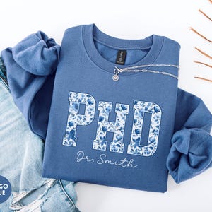 May include: Indigo blue sweatshirt with the letters "PHD" in a floral pattern. The name "Dr. Smith" is written below the letters in a cursive font. A silver necklace is draped across the sweatshirt. A pair of blue jeans is visible on the left side of the image.